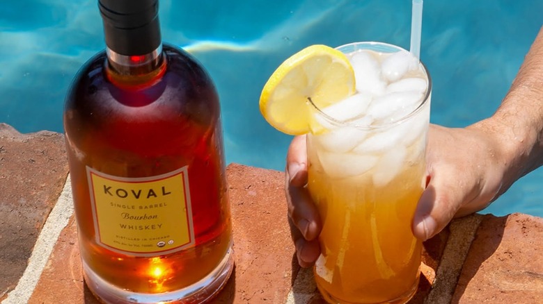 Bottle of KOVAL bourbon and hand holding cocktail on the edge of pool