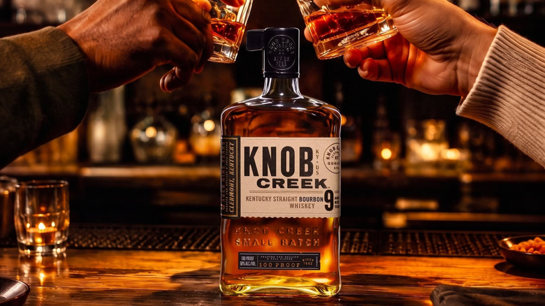 A bottle of Knob Creek 9-year placed on a bar with two people clinking glasses above it