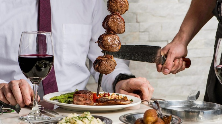 Skewer of meat being carved on a plate at a table set with sides and wine