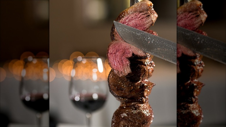 Steak being carved from a skewer with a red wine glass blurred in the background
