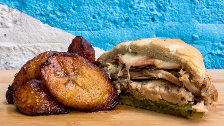 Plantains and a pork sandwich on a wooden table with a blue background