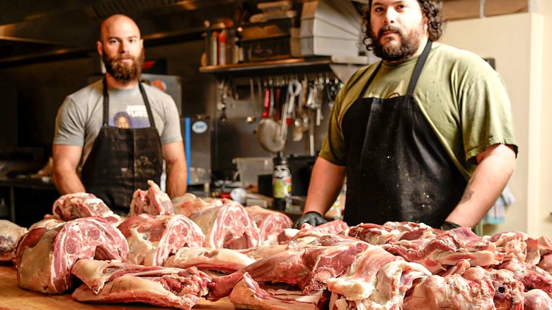 Two men standing behind butcher block full of fresh cut meats.