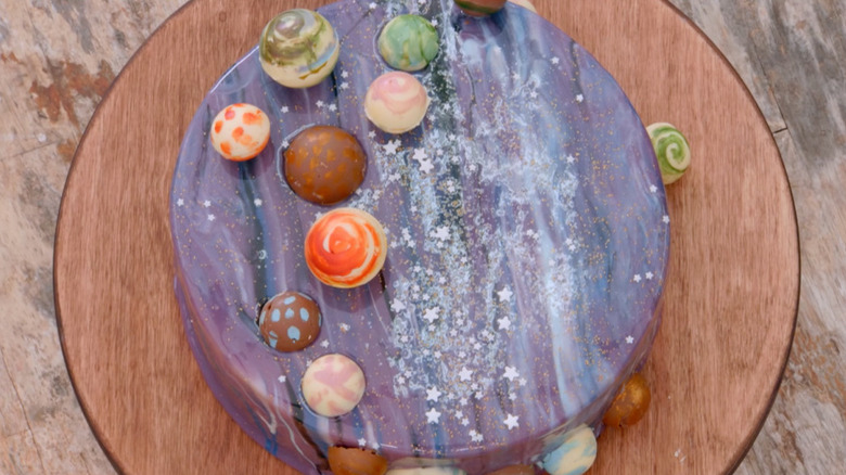 Cosmic cake with edible planets on a round, wooden cake stand on a table.