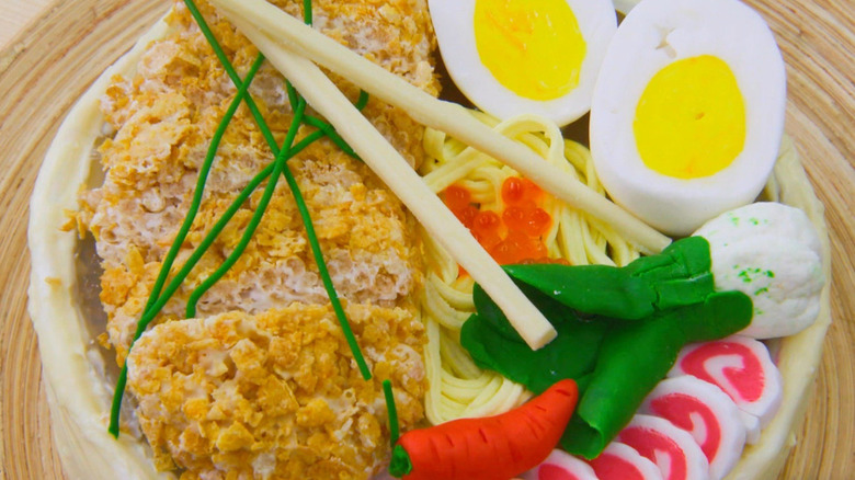A ramen shaped cake with eggs, noodles, vegetables and chopsticks on top of a bamboo placemat.