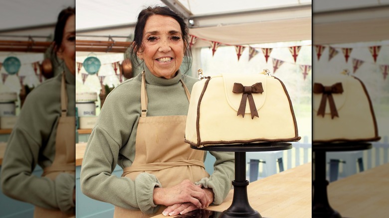 A purse shaped cake on a black pedestal with a woman in a green sweater and brown apron behind.