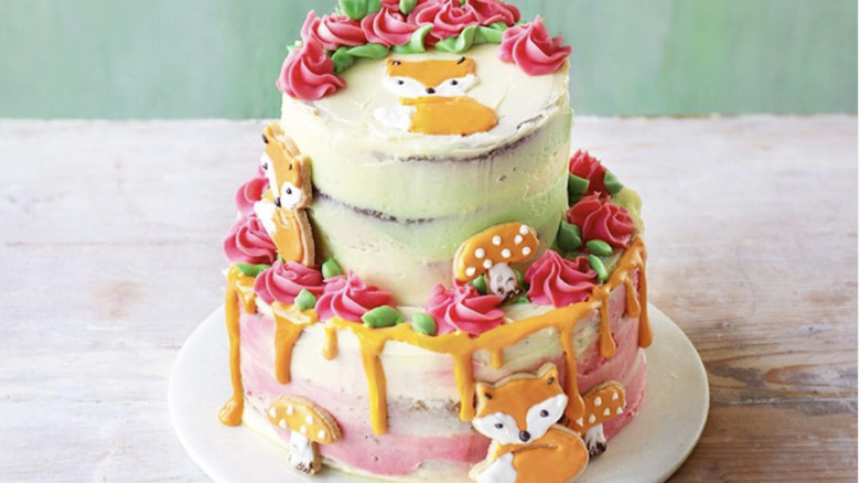 A two layer cake with pink flowers and foxes with dripping orange frosting on a white plate on a table.