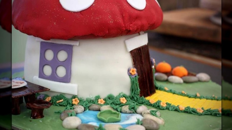 A toadstool house shaped cake on a bed of green frosting with candy pumpkins.