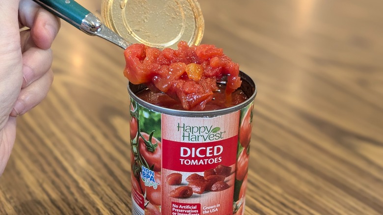 Hand spooning diced tomatoes out of open can with a wooden table in the back