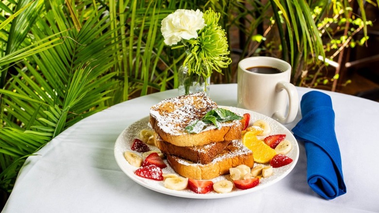 Plate of French toast with strawberries and bananas with coffee, flower, and plants in background