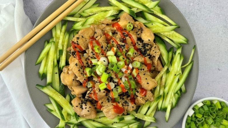 Bowl of bang bang chicken and cucumber batons, garnished with chopped green onions and hot sauce