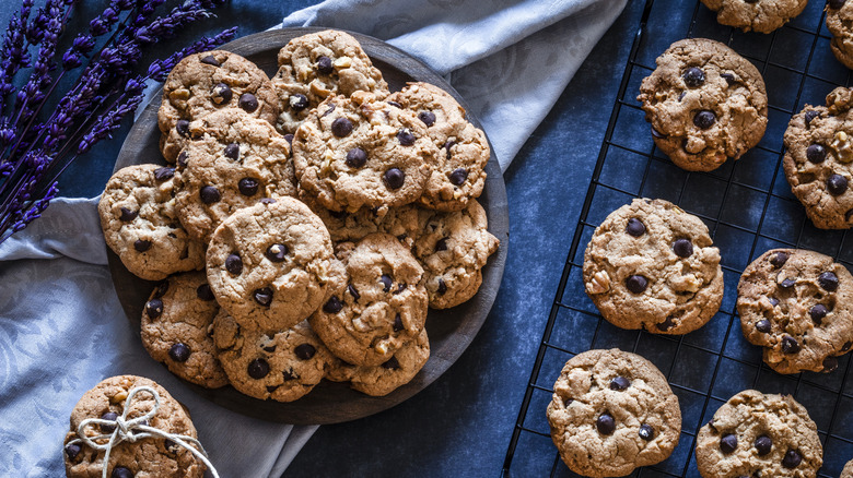 Chocolate chip cookies piled on a tin plate next a a baking rack with more cookies cooling, another bundle is tied with string. A sprig of lavender can be seen to the left