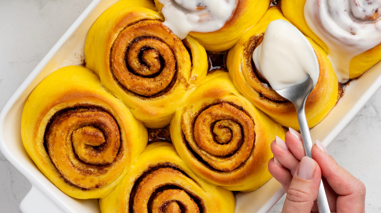 Spreading frosting over pumpkin cinnamon rolls in a baking dish with a spoon