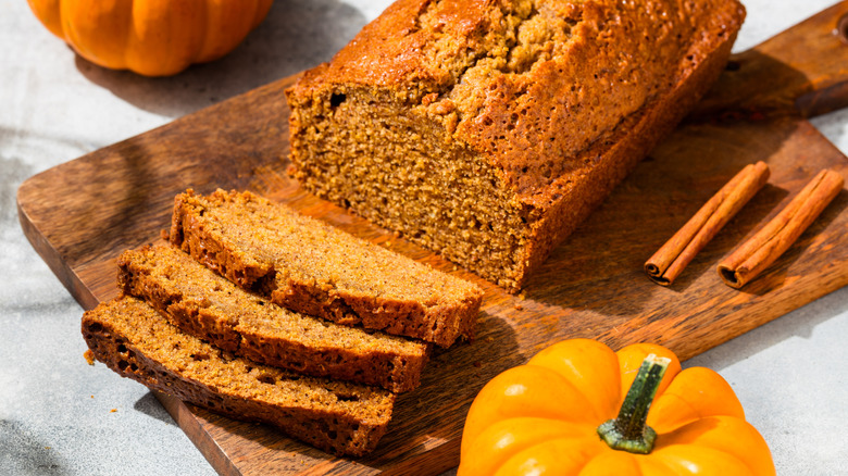 Sliced pumpkin bread on a wooden board next to cinnamon sticks and whole pumpkins