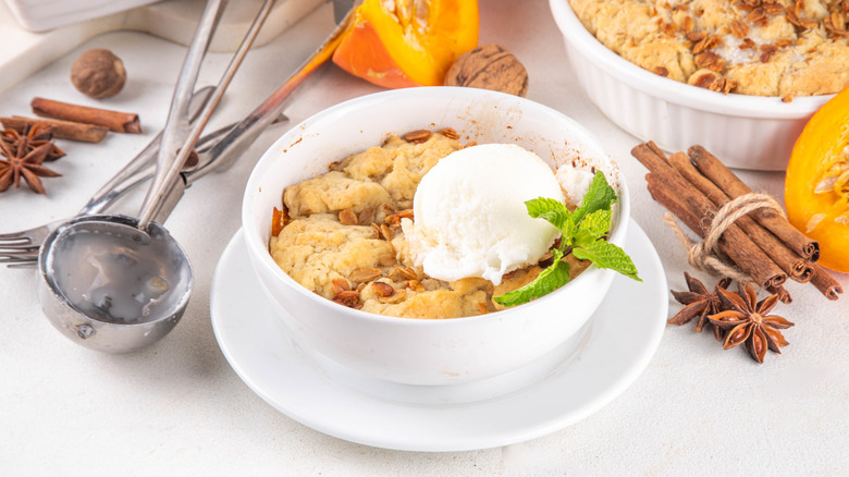 Bowl of pumpkin cobbler topped with a scoop of ice cream and a sprig of mint, next to whole spices and fresh pumpkin slices