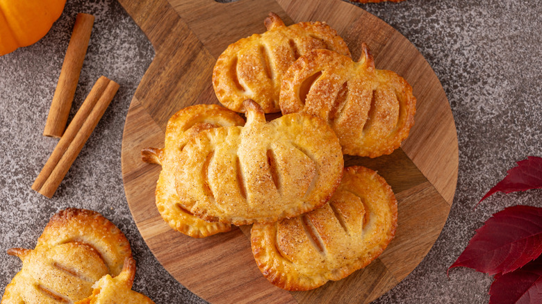 Pumpkin-shaped hand pies on a wooden serving board, next to cinnamon sticks and a pumpkin