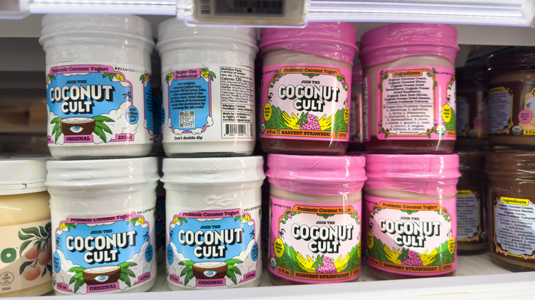 A jar of Coconut Cult Strawberry Harvest yogurt with a spoon in it against a wall