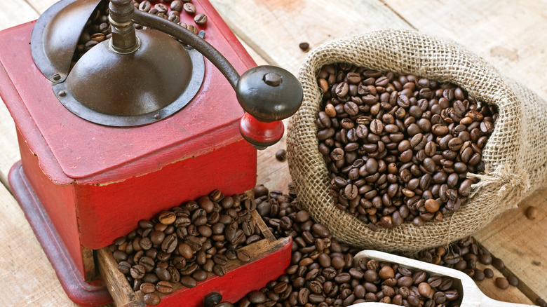 Whole bean coffee in a burlap sack next to an over-stuffed manual coffee grinder