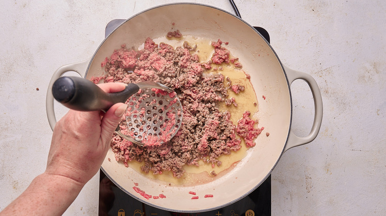 Aerial view of hands crumbling cooking beef with a potato masher in a pot