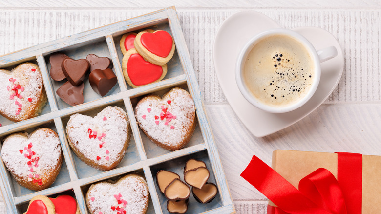 White box of heart-shaped cookies and chocolates next to white cup of coffee on heart shaped sauce and gift wrapped in red ribbon