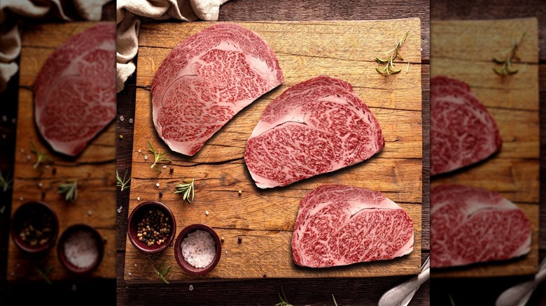 Three Wagyu beef ribeye steaks from Costco on wooden board next to rosemary springs and bowls of salt and pepper