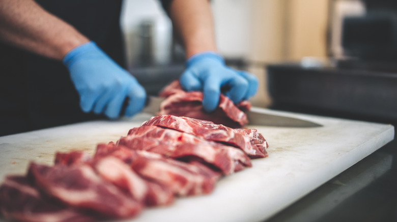 Someone cutting raw red meat on a plastic cutting board while wearing gloves