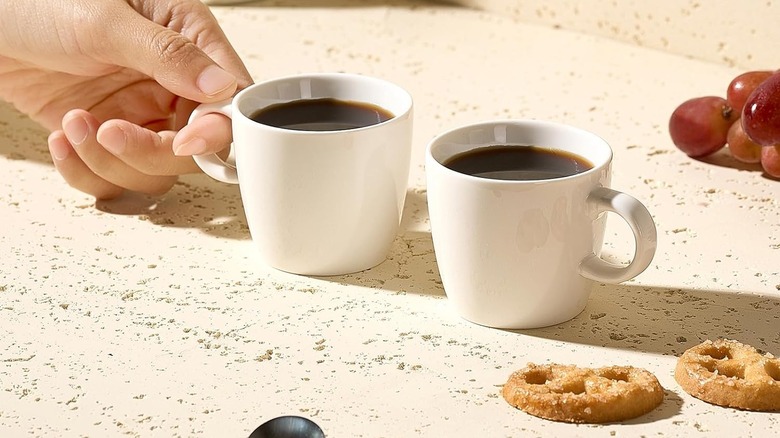 Two full Hlukana espresso cups on a countertop, with one held by a hand, alongside pretzels and grapes