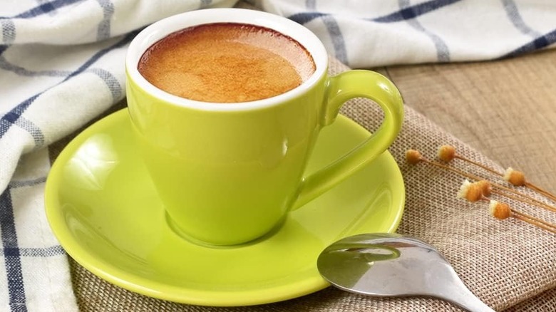 Close-up of a full ionEgg espresso mug on its saucer, with a cloth, spoon, and flowers in the background