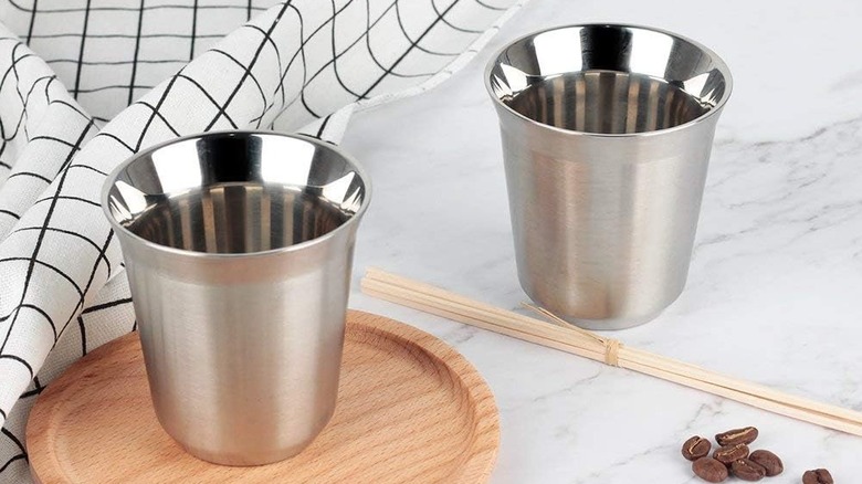 Two Recaps espresso cups on a kitchen surface with a cloth, wooden sticks, and coffee beans nearby