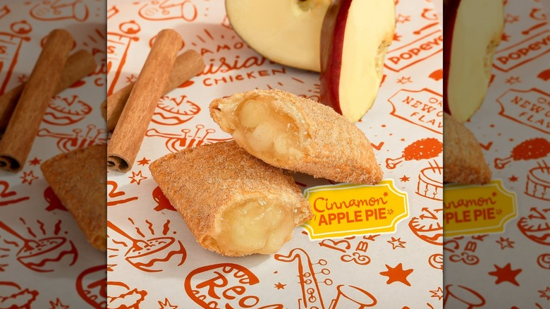 Halved Popeye's Cinnamon Apple Pie on branded background, next to cinnamon sticks and apple slices