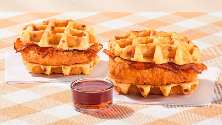 two Chick-fil-A chicken and waffles breakfast sandwiches sitting on napkins atop a checkered tablecloth with a small ramekin of syrup