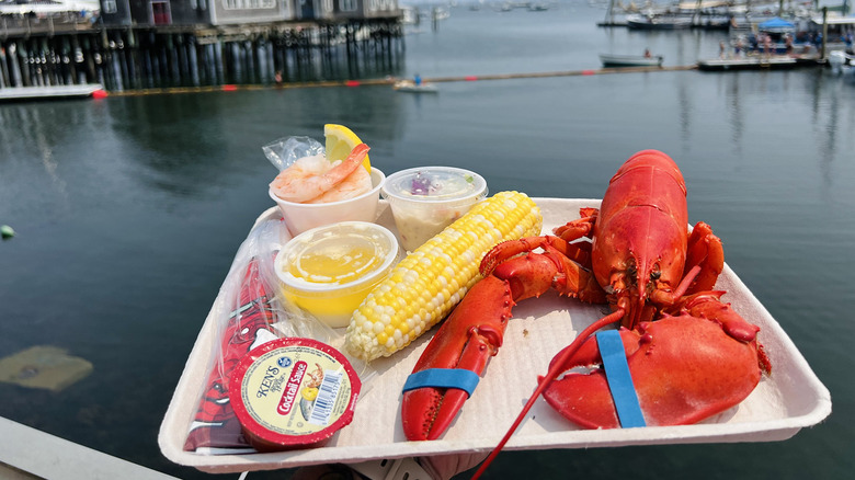 Hand holding a tray of lobster, corn cob, and shrimp by the water at the Maine Lobster Festival