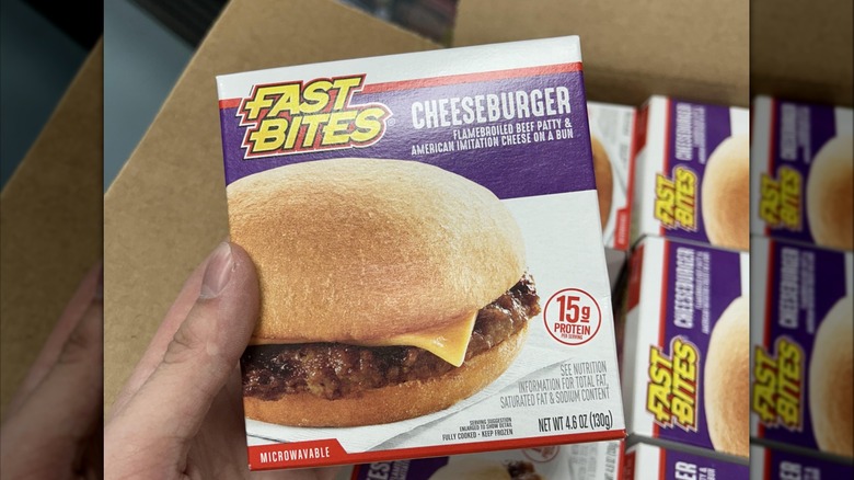 A hand holding a box of Fast Bites cheeseburgers over a case of the same