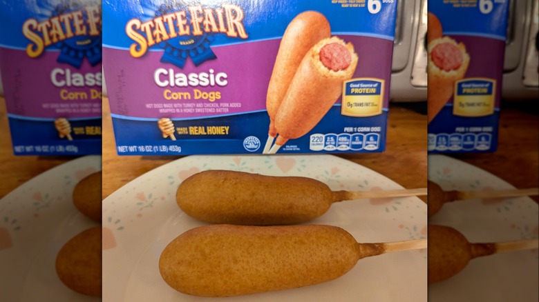A box of State Fair brand classic corn dogs sitting on a table behind a plate with two corn dogs on it