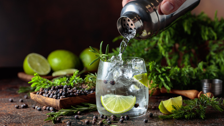 Hand pouring gin into a cocktail glass with limes and other garnishes in the background