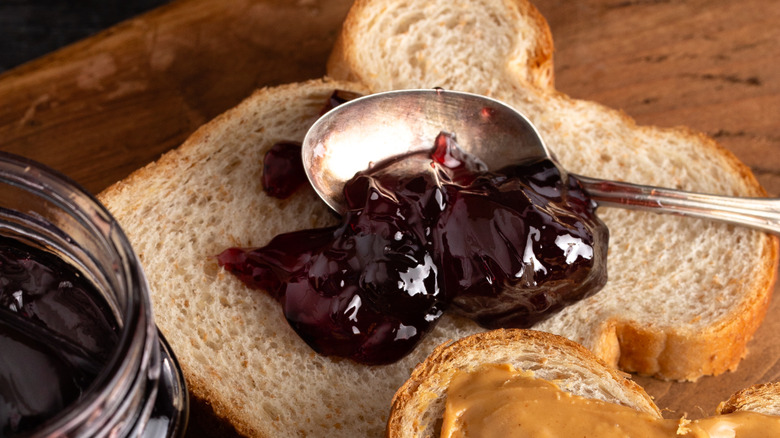 A wooden spoon of grape jelly next to a jar of the same and fresh grapes
