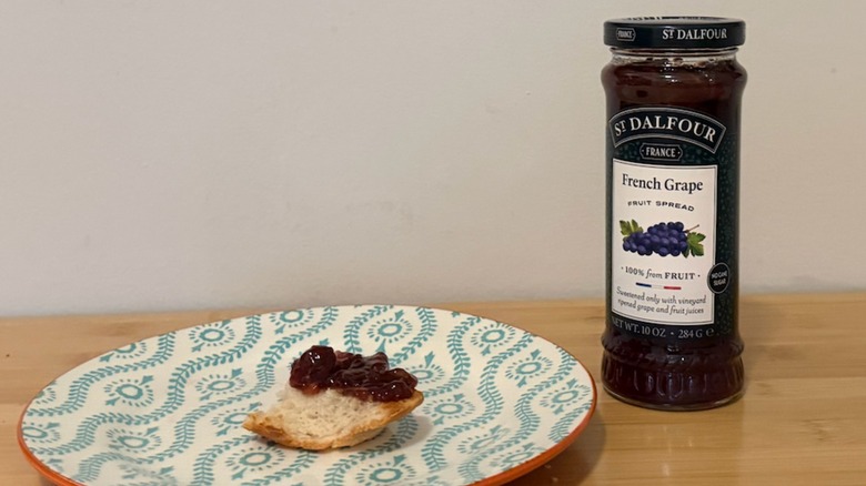 Small piece of jam-covered bread on a plate next to a jar of St. Dalfour French Grape Fruit Spread