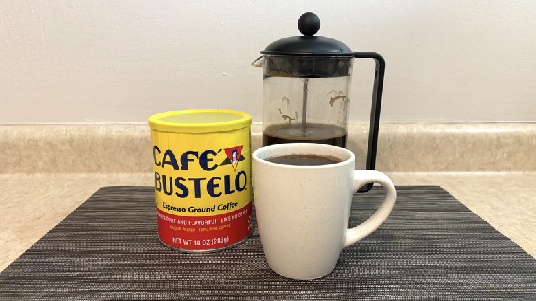 Café Bustelo in can, cup, and french press on black placemat
