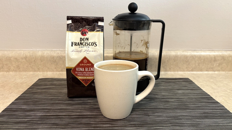 Don Francisco's Coffee Kona blend in bag, french press, and cup on black placemat
