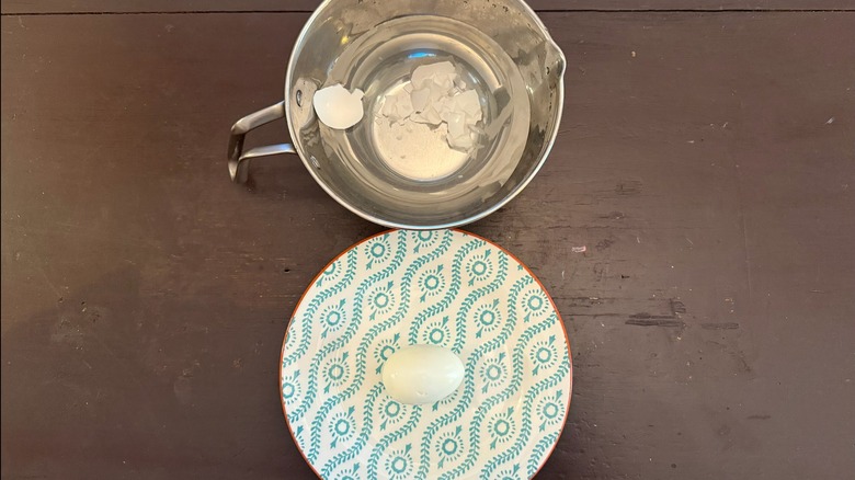 A peeled egg on a plate with a metal bowl of shells next to it