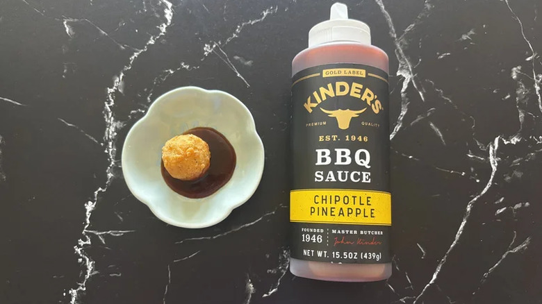 A bottle of Kinder's Chipotle Pineapple BBQ Sauce on a counter with a small dish of the sauce and a chicken nugget resting in it