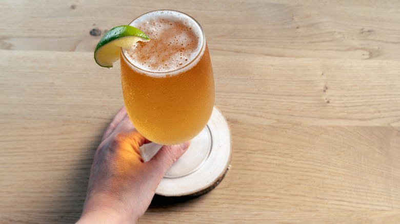 A close-up of a hand reaching for a glass of golden lager garnished with a fresh lime wedge