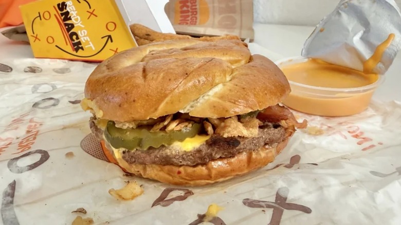 A Game Day Pretzel Burger sits in a box with a portion of unopened nacho cheese sauce and chicken fries tucked into the lid.