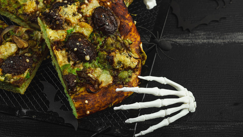 A skeleton hand and a spider reach for a green pizza on a black vinyl tablecloth.