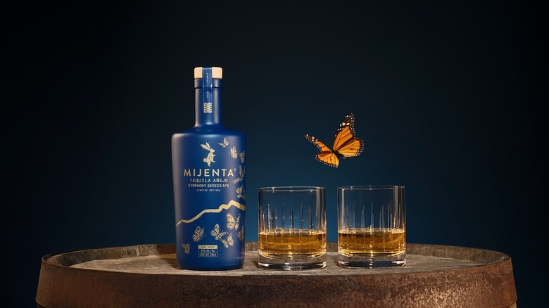 Blue bottle of Mijenta Tequila Añejo Symphony Series No 3 next to two glasses of tequila sitting on barrel, monarch butterfly beside