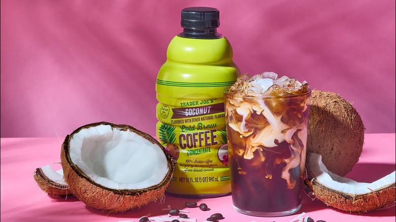 A photo of Trader Joe's Coconut Cold Brew Coffee Concentrate in a jug and over ice in a glass, surrounded by coconuts on a pink background