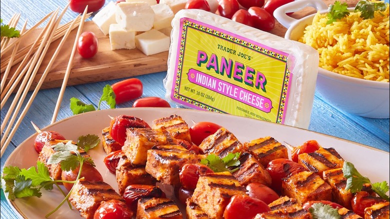 Trader Joe's Paneer Indian Style Cheese sitting on a blue wooden table along with a platter of kebabs, a bowl of rice, toothpicks, cubed cheese, and tomatoes