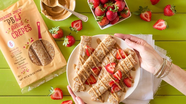 Trader Joe's PB&J Filled Crepes on a green wooden table, in a package and on a plate, with a bowl of strawberries and a bowl of peanut butter