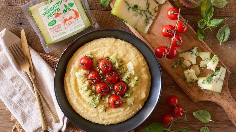 Trader Joe's The Sheepish Pesto in a package on a wooden table that has a cheeseboard with blue cheese, cherry tomatoes on the vine, a bowl of polenta with the same tomatoes on top, linen napkin, and silverware