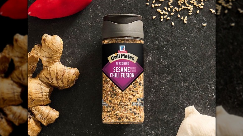 A jar of Grill Mates Sesame Chili Fusion seasoning on a countertop with ginger, garlic, red pepper, and sesame seeds nearby