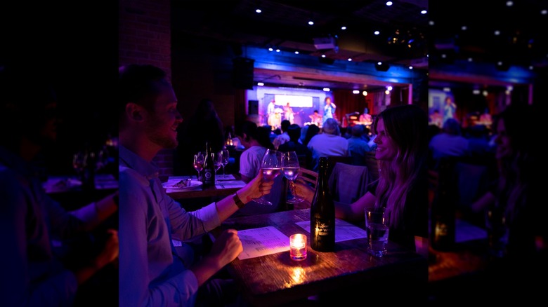 Two people toasting with wine glasses as live music plays on blurred stage in background